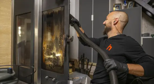 Un homme nettoie la vitre d'un poêle avec un aspirateur. Un bon entretien garantit la bonne fréquence de ramonage.