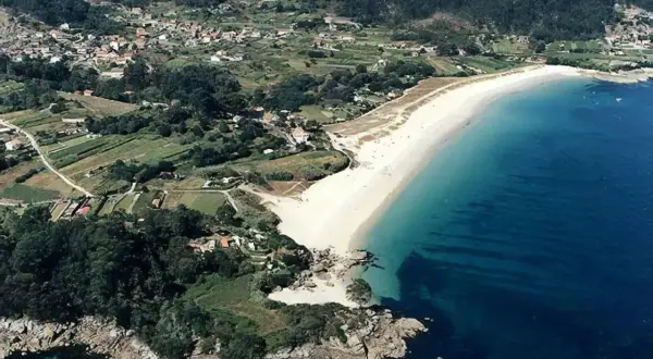 Vista aérea de la playa Area Brava, con arena blanca, aguas turquesas y un pueblo costero rodeado de vegetación y campos verdes.