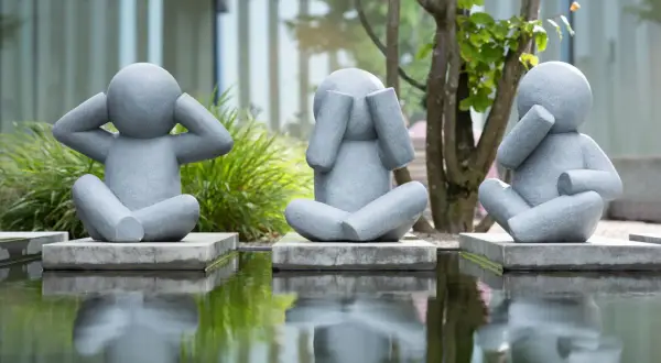 Drei graue Skulpturen von sitzenden Figuren, die ihre Ohren, Augen und Mund bedecken, spiegeln sich in einem Teich.