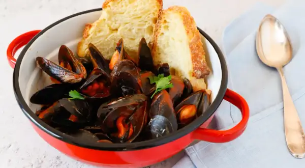 Un invitante guazzetto di cozze in una pentola rossa, accompagnato da pane croccante e una forchetta.