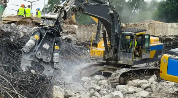 Maszyna budowlana wykonuje rozbiórkę, krusząc beton i stal. W tle widać pracowników w kamizelkach.