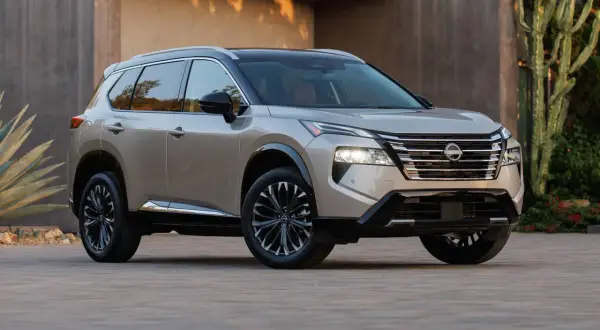 Elegancki, beżowy Nissan Rogue z czarnym dachem i przyciemnianymi felgami stoi przed nowoczesnym budynkiem, otoczony egzotyczną roślinnością.