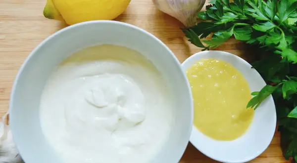 Zutaten für ein cremiges Aioli-Rezept: Knoblauch, Zitrone, Petersilie und zwei Schalen mit weißer und gelber Sauce.