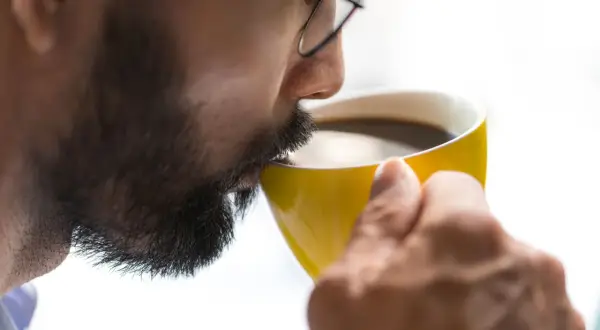 Man med glasögon och skägg dricker ur en gul mugg. Kanske känner han yrsel av kaffe.