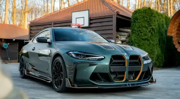 Matowy, zielony BMW M4 G82 z pomarańczowymi akcentami, zaparkowany przed drewnianym domkiem z boiskiem do koszykówki.
