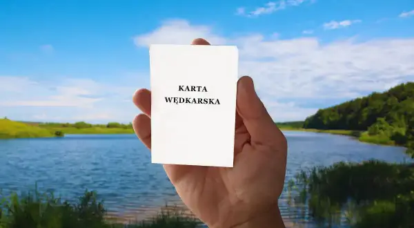Dłoń trzyma białą kartkę z napisem "KARTA WĘDKARSKA" na tle spokojnego jeziora i zielonych wzgórz.