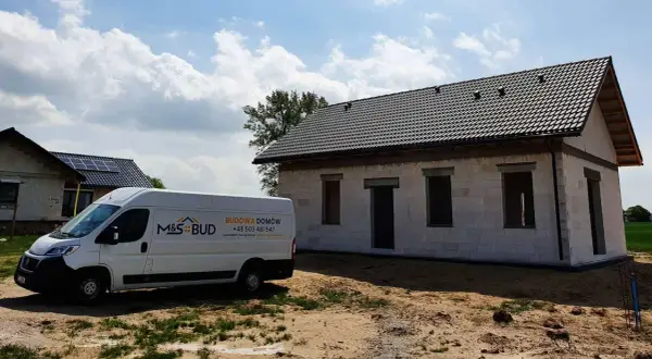 Biały van budowlany M&S BUD przed niedokończonym domem z szarej cegły pod czarnym dachem.