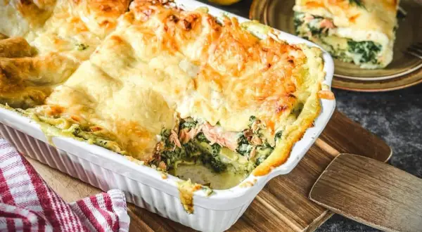 Herzhafte Spinat-Lachs-Lasagne, goldbraun überbacken, dampft in einer weißen Auflaufform. Ein Stück auf einem Teller daneben zeigt die Schichten.