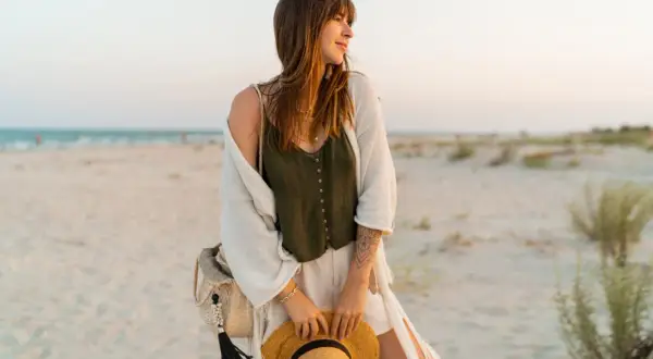 Kobieta w letnich stylizacjach boho jesień na plaży, z kapeluszem w dłoni i plecioną torbą.
