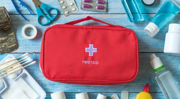 Czerwona apteczka z krzyżem i napisem FIRSTAID otoczona lekami, bandażami i akcesoriami medycznymi.