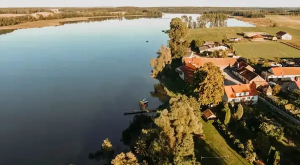 Spokojne jezioro Dejguny otoczone zielenią i domkami z czerwonymi dachami. W oddali widać lasy i pola.
