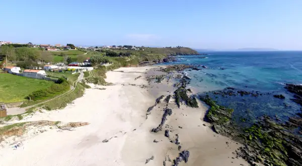 Vista aérea de la playa pragueira, con arena blanca, rocas cubiertas de algas y el mar azul turquesa. Casas y colinas verdes al fondo.