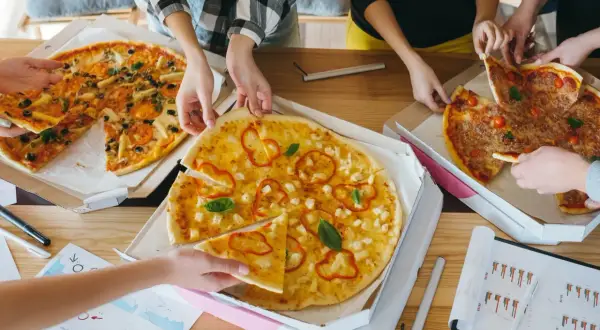 Kilka osób bierze kawałki pizzy z pudełek na drewnianym stole.