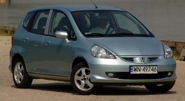 Jasnoszary Honda Jazz 2 z polskimi tablicami rejestracyjnymi WN 49746, zaparkowany przy wodzie.
