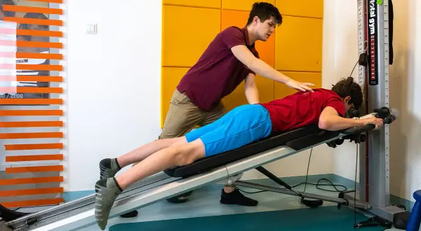 Fizjoterapeuta pomaga pacjentowi w ćwiczeniach na ławce rehabilitacyjnej. Skupiają się na rehabilitacji ortopedycznej, by odzyskać sprawność.