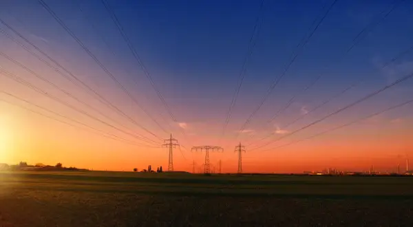 Linie energetyczne biegnące nad polem przy zachodzie słońca. W oddali widać przemysłowy krajobraz, przypominający o opłacie sieciowej stałej.