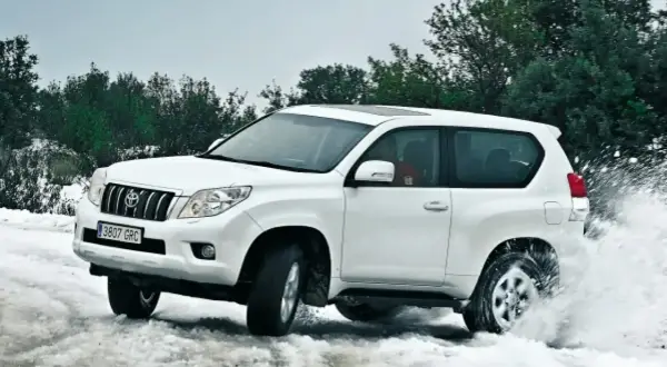 Toyota Land Cruiser 3.0 D4D 163 CV ficha técnica: un blanco todoterreno navega por nieve y agua, demostrando su potencia.