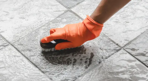 Mano con guanto arancione pulisce le fughe del pavimento con una spazzola, creando schiuma.