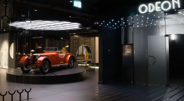 Pomarańczowy kabriolet z lat 20. XX wieku, symbol sztuki dwudziestolecia międzywojennego, prezentowany w nowoczesnym wnętrzu z neonowym napisem "ODEON".
