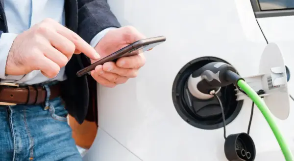 Mężczyzna sprawdza na telefonie, jak rozliczyć ładowanie samochodu elektrycznego w domu. Zielony kabel podłączony do pojazdu.