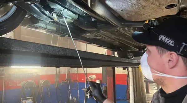 Mechanik w masce i rękawiczkach aplikuje środek konserwujący na podwozie samochodu. Długi aplikator pozwala dotrzeć do trudno dostępnych miejsc, co jest kluczowe, by wiedzieć, ile trwa konserwacja podwozia.