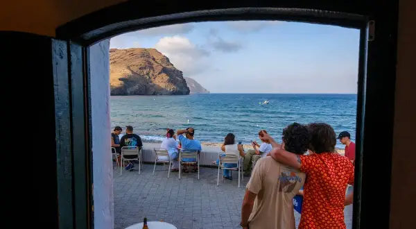 Dos amigos disfrutan de la vista al mar, con gente sentada en mesas. El manteca las negras, un día perfecto.