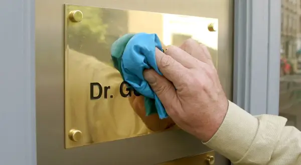 Hand reinigt Messing-Namensschild mit Tuch. Der Schriftzug "Dr. G." wird sichtbar.