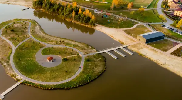 Malownicze, nisko jezioro z wyspą, ścieżkami, altanką i nowoczesnym budynkiem. W tle widać drzewa, plażę i zabudowania.