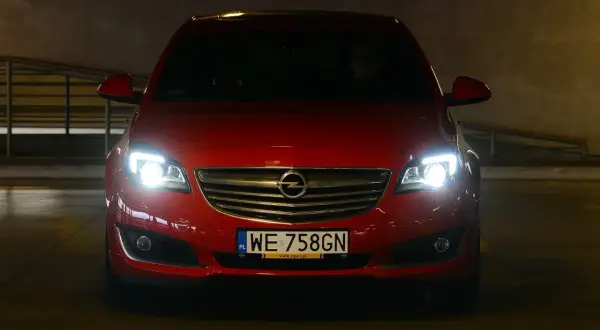 Czerwony Opel z włączonymi światłami, gotowy do jazdy. Dowiedz się, jak ustawić światła w samochodzie domowym sposobem, by zapewnić sobie bezpieczeństwo.