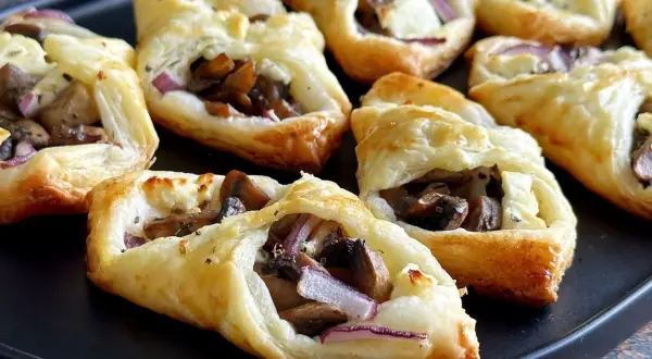 Chrupiące ciasto francuskie z pieczarkami, serem feta i czerwoną cebulą, ułożone na czarnej tacy.
