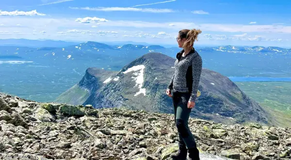 En kvinna står på en stenig topp med utsikt över ett vidsträckt fjällandskap. En vy som liknar den från en **webbkamera Helags**.