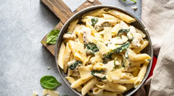 Penne arrabiata z kurczakiem i szpinakiem, posypane parmezanem, podane w patelni na drewnianej desce.