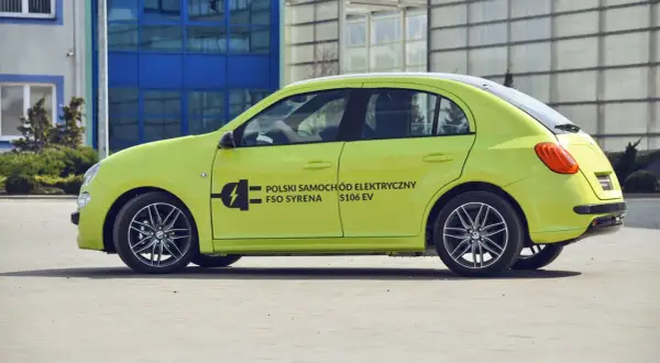 Jasnozielony FSO Syrena S106 EV, jeden z polskich samochodów elektrycznych, parkuje przed nowoczesnym budynkiem.