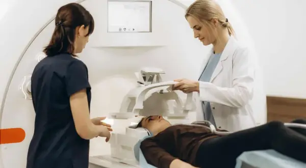 Zwei medizinische Fachkräfte bereiten eine Patientin für radiologische Untersuchungen vor. Sie liegt auf einer Liege neben einem MRT-Gerät.