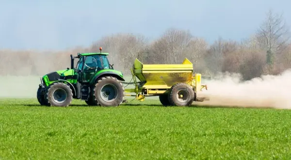 Tractor verde esparciendo cal viva en la huerta, creando una nube de polvo sobre el campo verde.