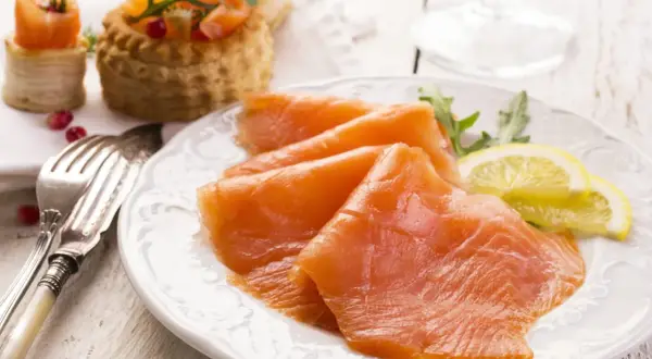 Tranches de saumon fumé, garnies de roquette et de citron, sur une assiette blanche. Des bouchées feuilletées au saumon sont visibles en arrière-plan.