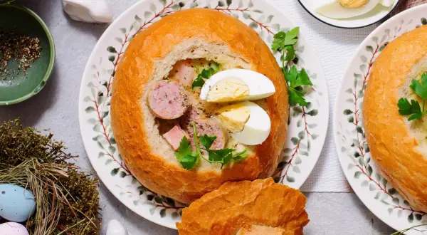 Pyszny żur w chlebie z kiełbasą i jajkiem, ozdobiony natką pietruszki. Na stole wielkanocne dekoracje: pisanki w gnieździe, zajączek.