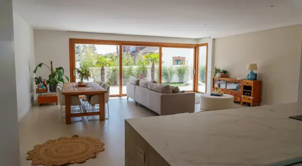 Salón moderno con comedor y sofá, destacando una pared prefabricada interior que se integra con grandes ventanales al jardín.