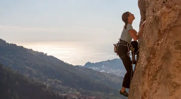 Eine Frau klettert an einer Felswand in Finale Ligure. Im Hintergrund glitzert das Meer.