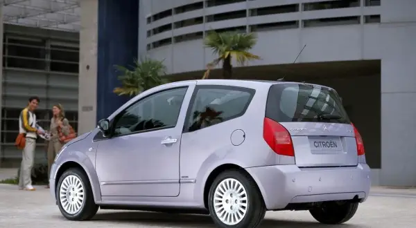 Jasnoszary Citroën C2 z parą stojącą w tle przed nowoczesnym budynkiem.