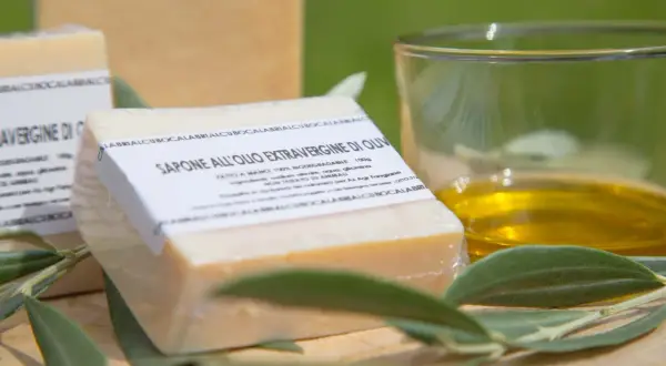Sapone all'olio extravergine di oliva, artigianale e biodegradabile, accanto a un bicchiere d'olio e foglie d'ulivo.