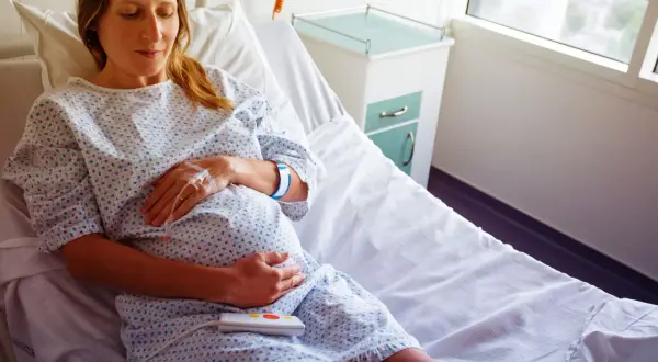 Kobieta w szpitalnym łóżku, z kroplówką i opaską na ręce, czeka na dziecko. Jej tryb życia po założeniu szwu szyjkowego jest spokojny.