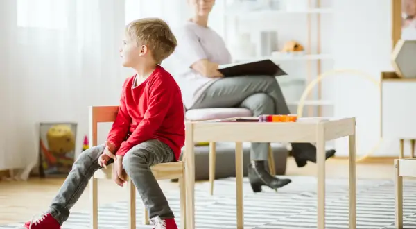 Pojke i röd tröja sitter på en stol under en autism utredning barn. En kvinna sitter bakom ett bord.