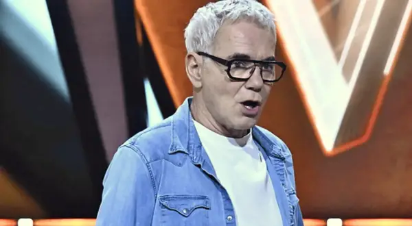 Czy program Jaka to melodia zostanie skasowany z powodu spadku oglądalności? Hubert Urbański w okularach i jeansowej koszuli na tle scenografii.