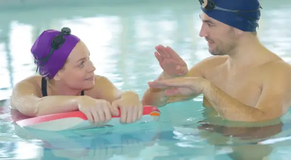A man and woman in a pool, likely during adult swim lessons, with the man demonstrating a swimming technique.