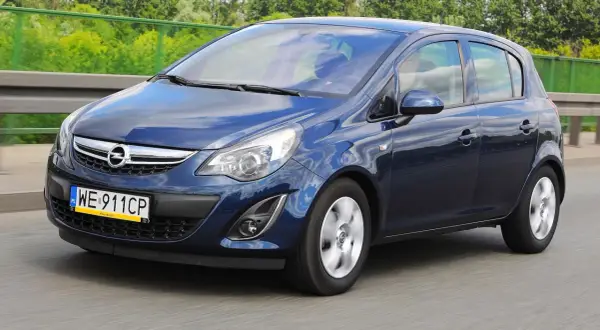Granatowy Opel Corsa D jedzie po drodze, odbijając niebo i chmury.