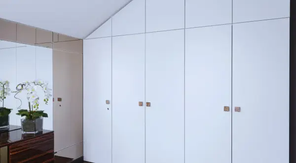 Armario empotrado con puertas blancas y tiradores cuadrados. Reflejos de orquídeas y un mueble de madera oscura en el espejo. Ideal para cambiar puertas armario empotrado.