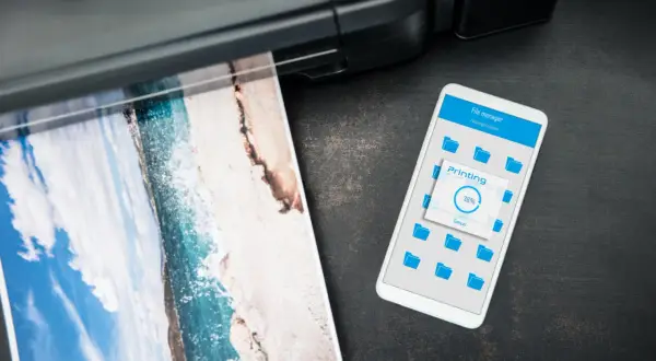 Jak wydrukowac cos z iPhone? Widok telefonu z ekranem "Printing 38%" obok drukarki drukującej zdjęcie plaży.