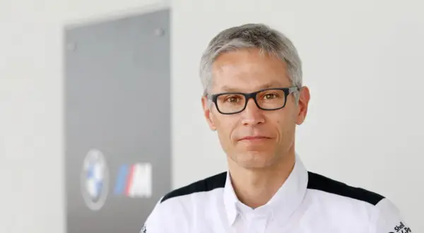 Mike Krack, con gafas y camisa blanca con detalles negros, posa frente a un logo de BMW M.