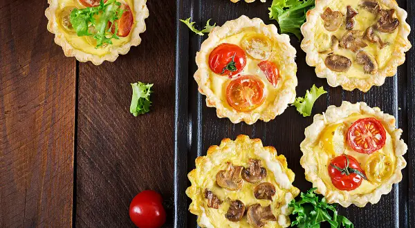 Mini quiches apéro aux tomates et champignons, garnies de feuilles de salade verte, sur fond de bois sombre.